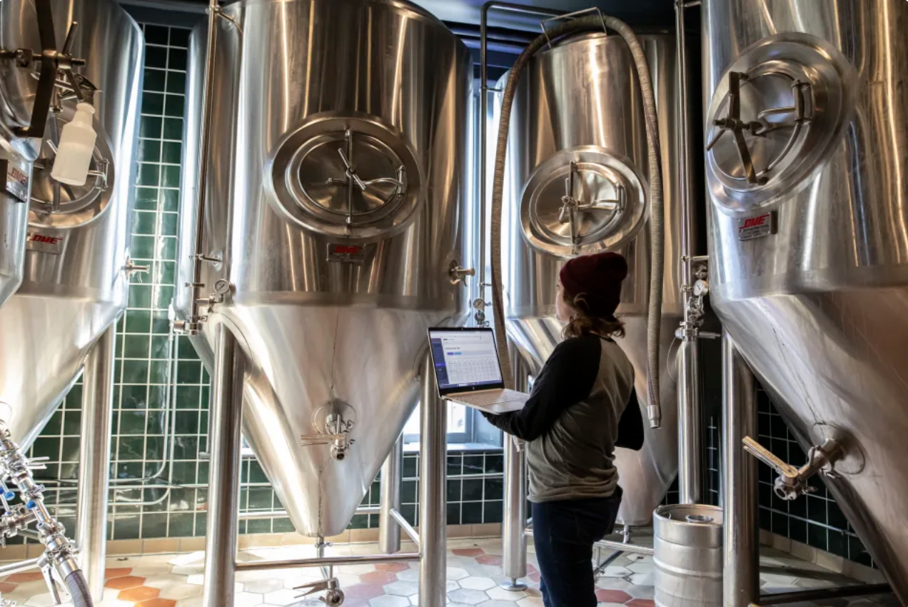 Brewer holding a laptop while standing in front of beer tanks. MarginEdge + Arryved integration